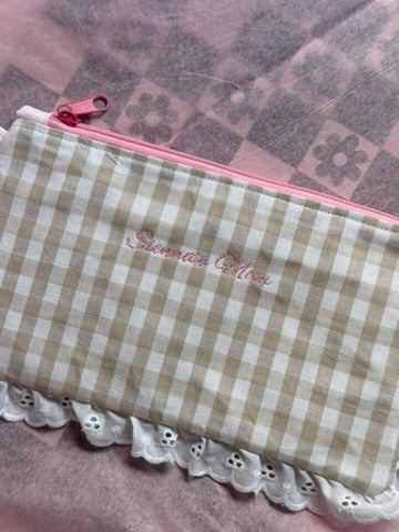 A beige and white gingham check pencil case with a pink zipper and a white eyelet lace trim along the bottom edge.  'Sienna's Mom' is embroidered in pink script on the front.