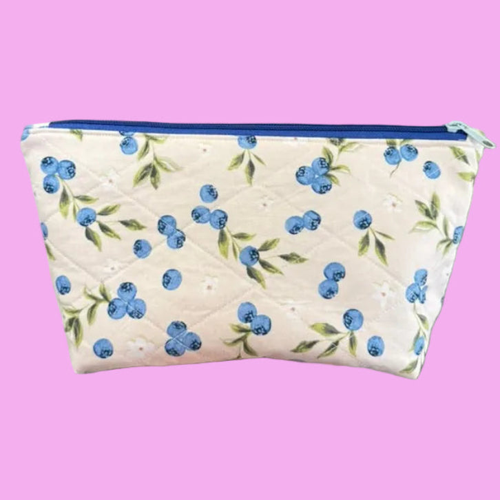 A hand holding a quilted cosmetic pouch with a blueberry pattern. The pouch is off-white with blue and green blueberry prints and a blue zipper.