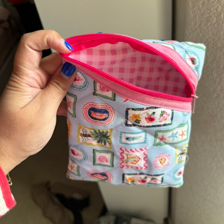 A hand holding an open zippered pouch with a light blue and pink gingham lining and a colorful patchwork-style print on the exterior.