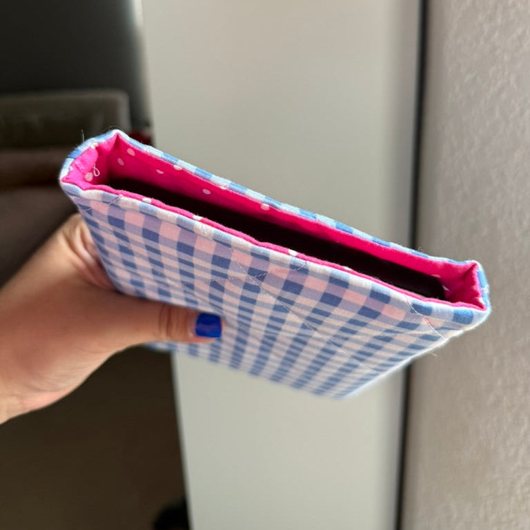 A hand holding a small fabric pouch or sleeve with a blue and white gingham pattern and a bright pink polka-dot interior lining, shown against an indoor background.