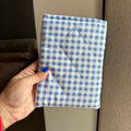 A hand with blue nail polish holds a small, square, quilted fabric pouch or case featuring a blue and white gingham checkered pattern.