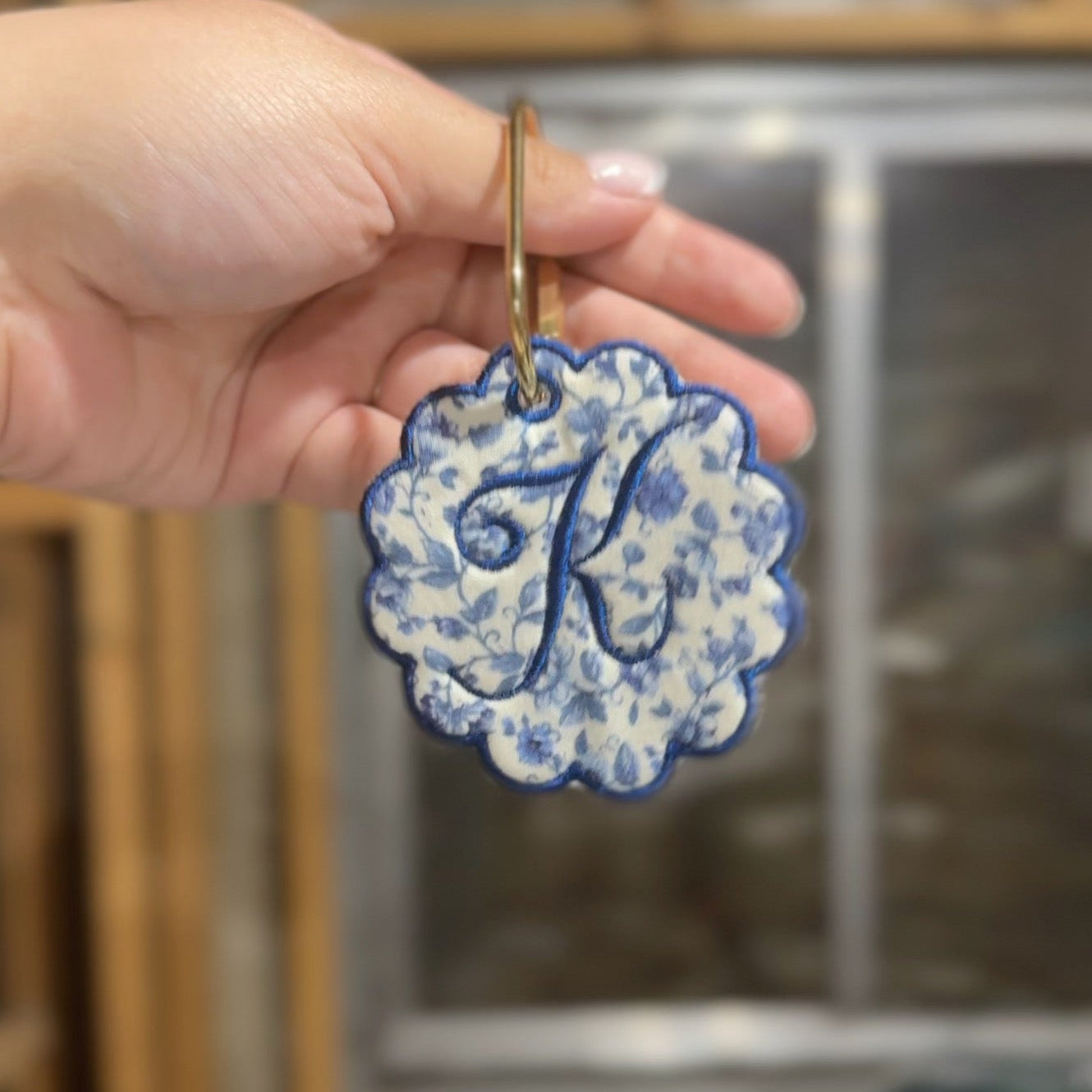 Decorative keychain with floral pattern and letter 'K' held by a hand against a blurred background.