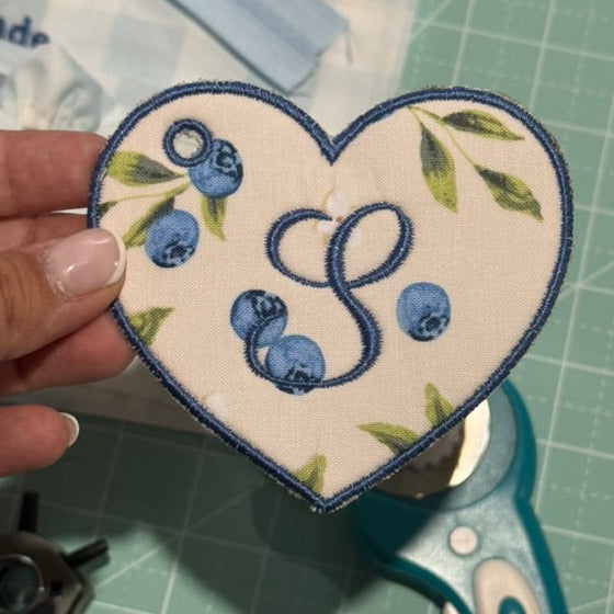 Heart-shaped embroidered patch with blueberries and 'S' initial held over a cutting mat with scissors.