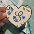 Heart-shaped embroidered patch with blueberries and 'S' initial held over a cutting mat with scissors.