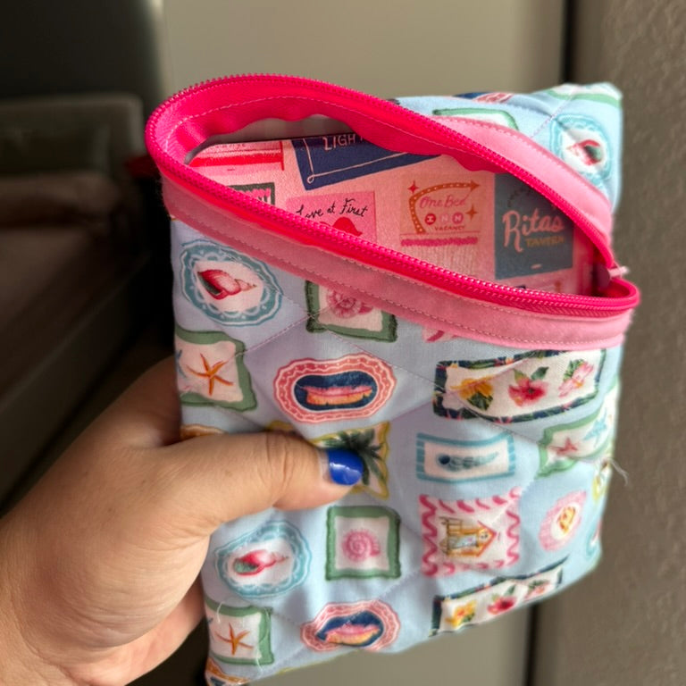 A hand holding a small zippered pouch with a light blue and pink patchwork print, showing a colorful interior lining.