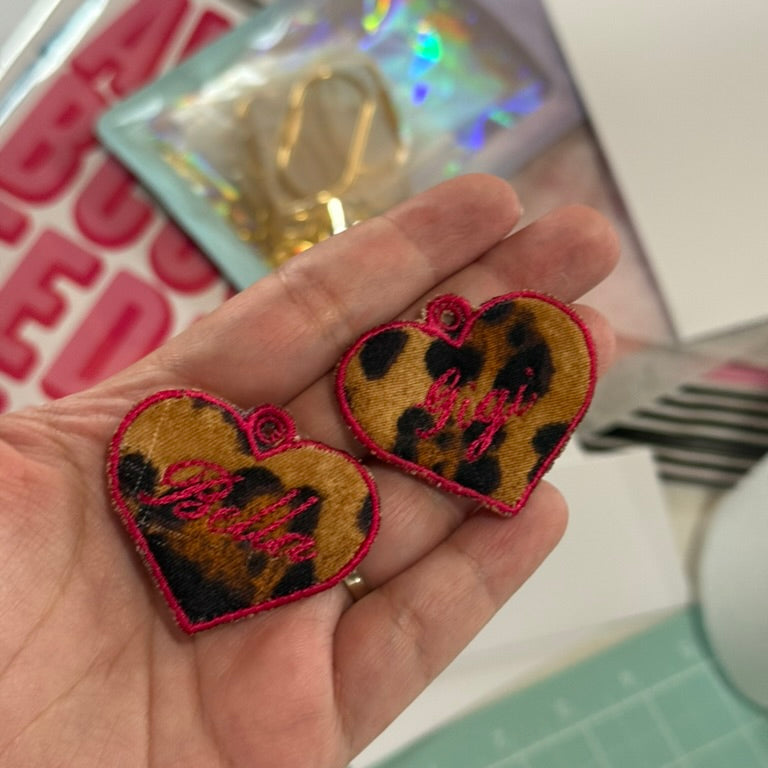 Heart-shaped patches with leopard print and red text held in a hand, with a blurred background.