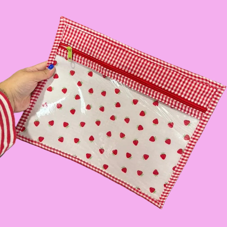 A hand holds a clear zippered pouch with a red and white gingham border and a red strawberry print on a white background.