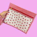A hand holds a clear zippered pouch with a red and white gingham border and a red strawberry print on a white background.