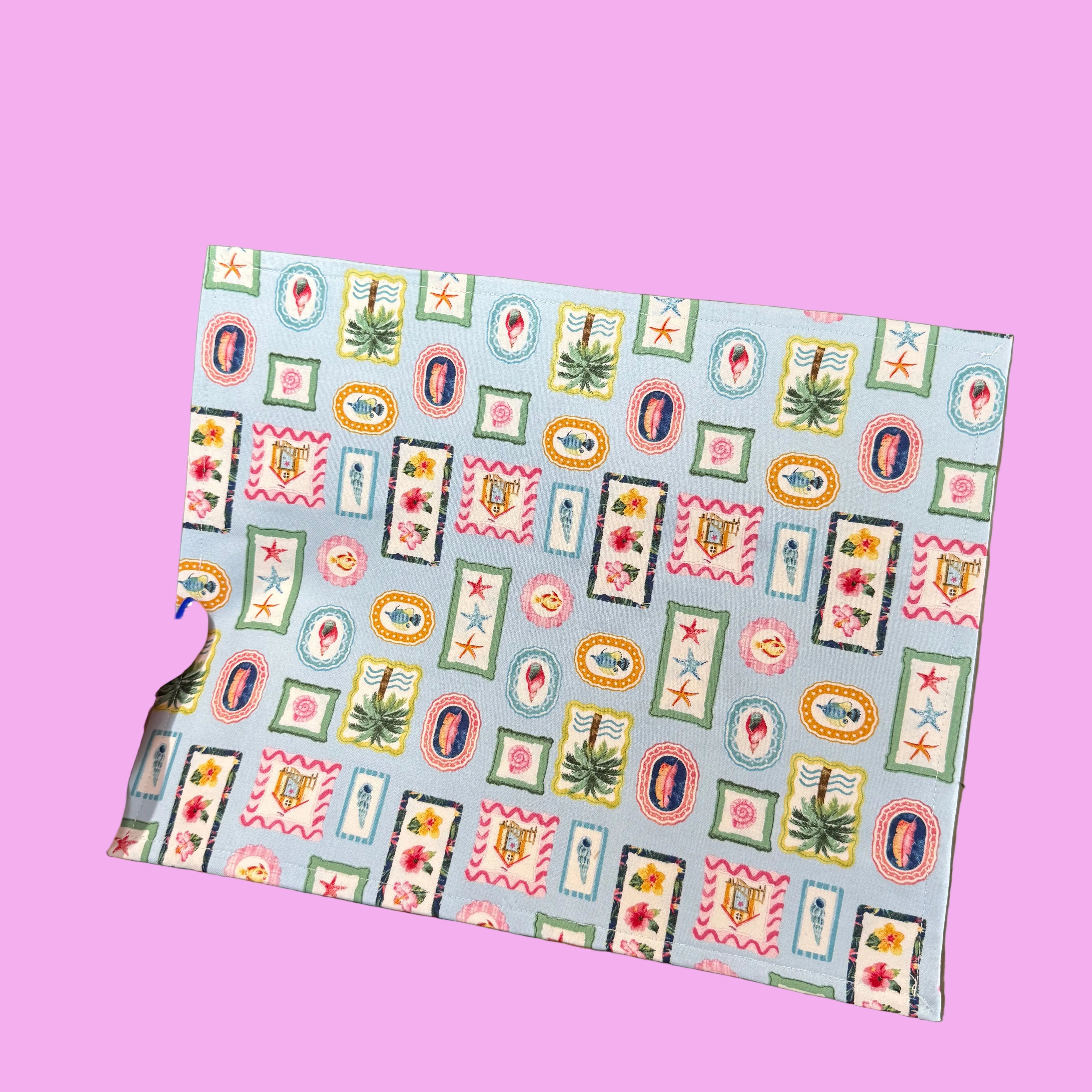 A hand with blue nail polish holds up a square piece of fabric featuring a colorful patchwork pattern of vintage stamps, stars, and floral motifs on a light blue background. 