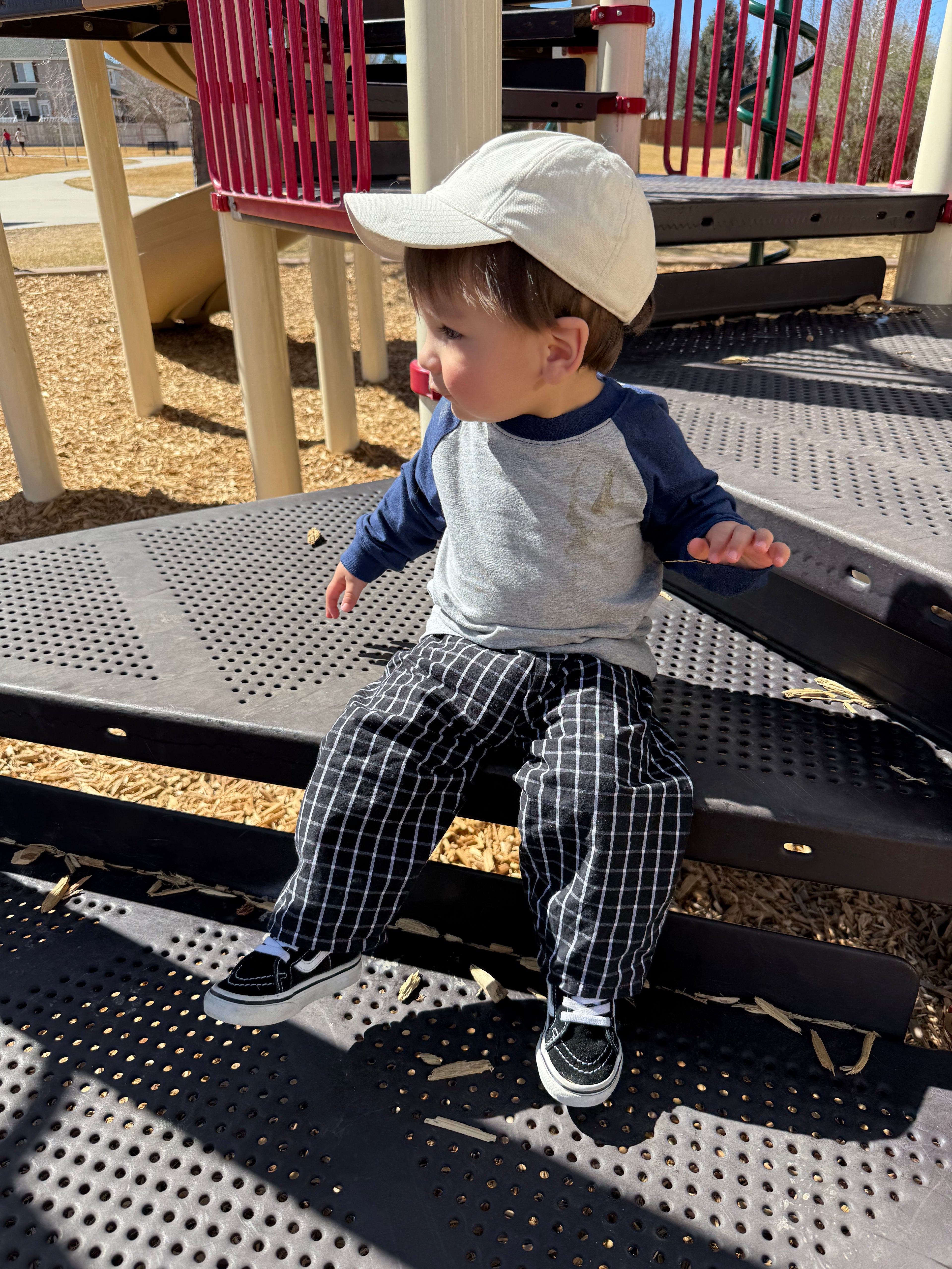 Child sitting on a playground bench wearing a cap and checkered pants.