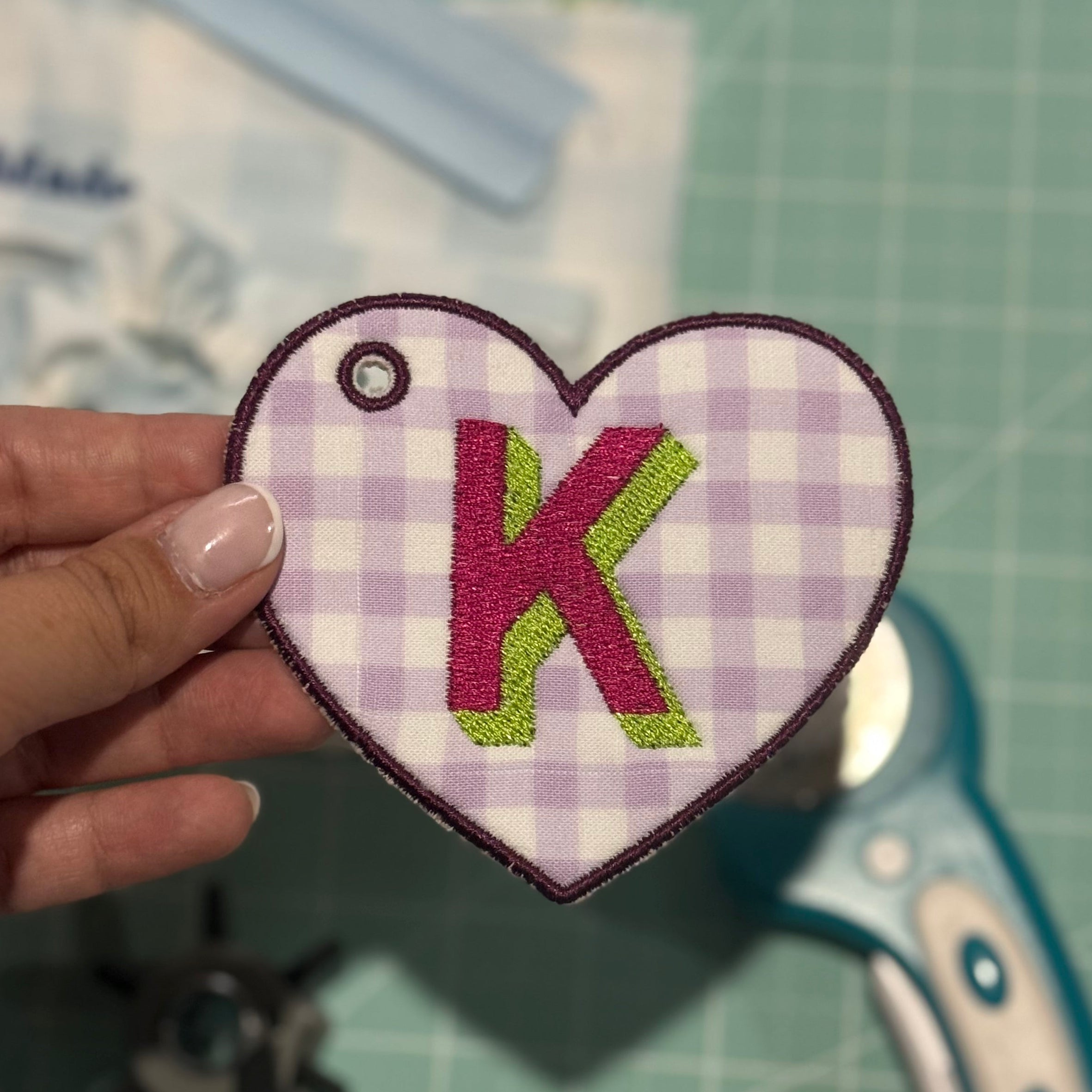 Heart-shaped embroidered patch with letter 'K' held over a cutting mat with scissors.